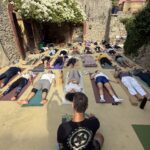 Turma em śavāsana no pátio, sob as buganvílias brancas; Pedro, de costas, observa